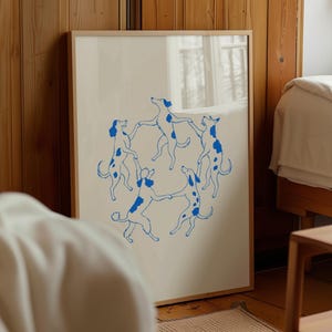Puede incluir: Una lámina de arte enmarcada que presenta una ilustración circular de cinco perros azules con patrones de manchas que se sujetan de las patas en un círculo. La obra está sobre un fondo blanco roto y en un marco de madera de color claro.