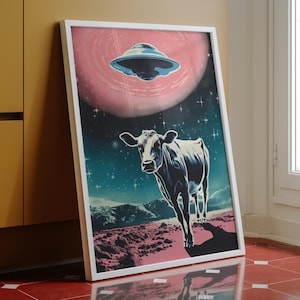 May include: A framed wall art print featuring a surreal scene of a cow standing on a rocky, pink-toned landscape under a dark starry sky. Above the cow, a metallic flying saucer hovers against a large, circular, glowing pink planet or moon.