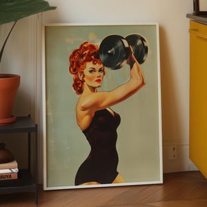 Vintage 1950s style pin-up girl art print featuring a strong woman with red hair lifting a heavy black dumbbell overhead. Retro fitness wall art showing a muscular woman in a black swimsuit, perfect for a vintage home gym decor, garage art, or mid-century modern aesthetic.