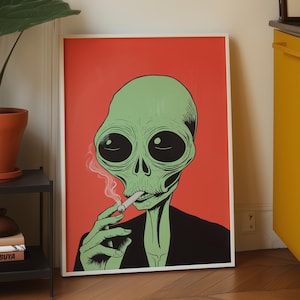 May include: A framed rectangular wall art print featuring a green alien with large black eyes wearing a black shirt, smoking a cigarette with visible pink smoke, set against a solid red background, displayed in a white frame.