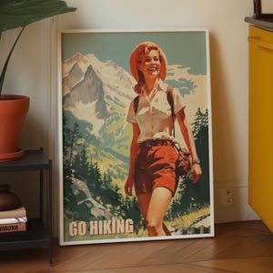 May include: A framed vintage poster depicting a woman hiking in the mountains. The poster shows a woman in a white shirt, red shorts, and a hat, with the words "GO HIKING" at the bottom. The background features a mountain range and trees.