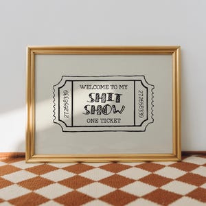 May include: A framed print with a gold-colored frame. The print features a ticket design with the words "WELCOME TO MY SHIT SHOW ONE TICKET" in black. The background is a light beige color.