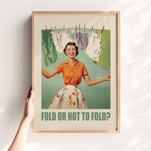 May include: A framed poster featuring a smiling person in an orange button-down shirt and a floral skirt standing before a clothesline with hanging laundry. The text FOLD OR NOT TO FOLD? is printed in green at the bottom of the cream-colored background.