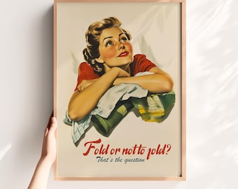May include: A framed poster featuring a vintage-style illustration of a person with brown hair wearing a red top, leaning on a pile of folded laundry. The text reads, Fold or not to fold? That's the question, in red and dark blue lettering.