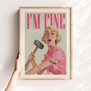 May include: Framed vintage-style print with the text "I'M FINE" in pink letters. The artwork depicts a woman in a pink shirt holding a hammer with an expression of shock. The print is in a wooden frame.