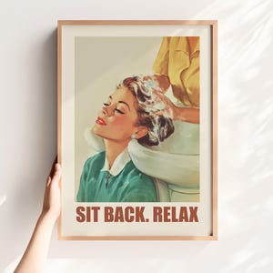 May include: A framed vintage-style print featuring a woman getting her hair washed in a salon. The woman is wearing a green top and has her eyes closed. The text "SIT BACK. RELAX" is printed in brown at the bottom of the print.
