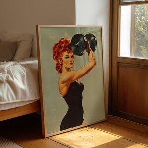 Vintage 1950s style pin-up girl art print featuring a strong woman with red hair lifting a heavy black dumbbell overhead. Retro fitness wall art showing a muscular woman in a black swimsuit, perfect for a vintage home gym decor, garage art, or mid-century modern aesthetic.