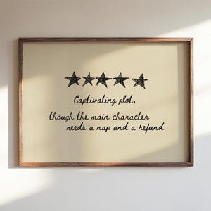 May include: A framed print with a light beige background and a brown wooden frame. The print features five black star illustrations above handwritten text that reads, "Captivating plot, though the main character needs a nap and a refund."