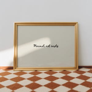May include: A gold-framed picture with the handwritten text "Minimal, not empty" on a light-colored background. The frame is rectangular and sits on a brown and white checkered surface.