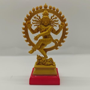 May include: A golden statue of the Hindu deity Shiva Nataraja, the dancing god. The statue features Shiva in a dynamic pose within a circular frame, atop a small figure. The base is red, and the overall design is intricate.