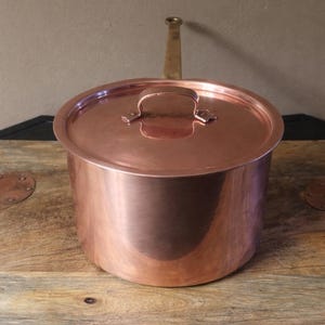 May include: A copper pot with a lid and a brass handle. The pot is cylindrical with a rounded lid and a small handle on top. The pot sits on a wooden surface.