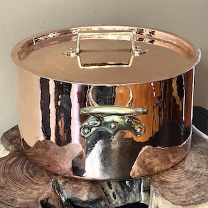 May include: A polished copper stockpot with a matching lid. The pot features two side handles made of brass, each secured with three rivets. The lid has a central rectangular brass handle. The exterior surface is reflective and smooth.