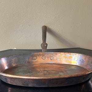 May include: An oval copper pan with a weathered, multi-toned metallic finish featuring a dark, oxidized patina. The pan includes a single, sturdy metal handle attached to the rim with three visible rivets, showcasing a worn, industrial aesthetic.