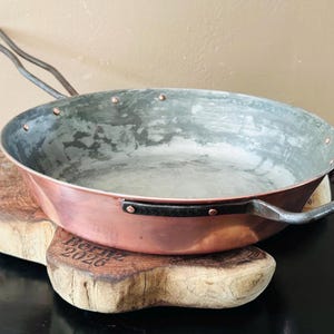 May include: A round copper pan with a metallic silver-toned interior lining, featuring a long, curved iron handle and a smaller side handle. The pan is secured with copper rivets along the rim and is resting on a natural wood slab with the text Bernz 2026.