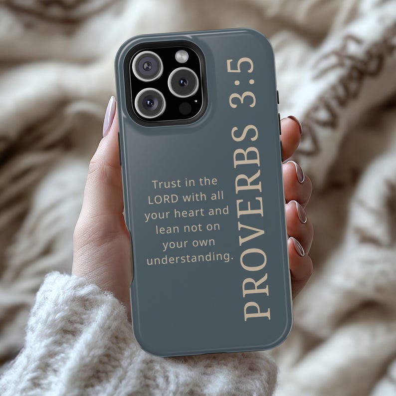 May include: A slate-gray phone case with the text "PROVERBS 3:5" in a vertical orientation. The case also features the quote "Trust in the LORD with all your heart and lean not on your own understanding." The phone case has a camera module with three lenses.