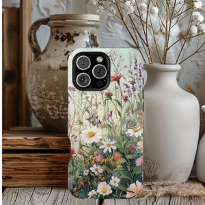 May include: A smartphone case featuring a printed design of a wildflower meadow with white daisies, pink blossoms, and purple sprigs on a pale green background. The case has a black camera cutout section with three circular lenses and a flash.