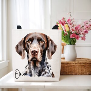 May include: A canvas tote bag featuring a watercolor portrait of a German Shorthaired Pointer dog. The dog has brown and white markings, with the name "Orion" written in script. The bag has black handles.