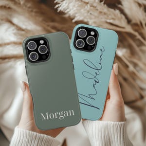 May include: Two phone cases, one sage green and one teal, are held up. The sage green case has the name "Morgan" printed on it. The teal case has the name "Madeline" written in cursive. Both cases have a camera array.