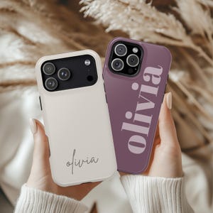 May include: Two phone cases, one cream-colored and one purple. The cream case has the name "Olivia" in a cursive font. The purple case has the name "Olivia" in a bold, sans-serif font. Both cases have a camera cutout.