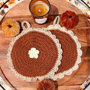 May include: Two round, crocheted pot holders in shades of brown and cream, each with a small white flower in the center. A lit candle in a glass jar and several small orange velvet pumpkins are arranged around the pot holders.