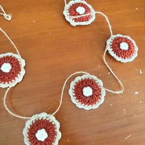 May include: A handmade crochet garland featuring flower-shaped ornaments. Each flower has a dark red center, a cream-colored border, and a white center. The flowers are strung together with natural twine, creating a rustic and charming decorative piece.