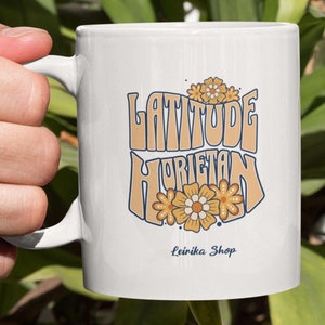 Może przedstawiać: Biały ceramiczny kubek z napisem "LATITUDE HORIZON" w kolorze pomarańczowym i niebieskim, z kwiatowymi akcentami. Kubek ma również napis "Leinika Shop". Kubek jest trzymany w dłoni.