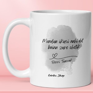 May include: White ceramic mug with a handle. The mug features a watercolor-style gray blotch with black script that reads "Mundua ikusi nahi dut baina zure ohetik" and "Berri Txarrak". A small heart is also included.
