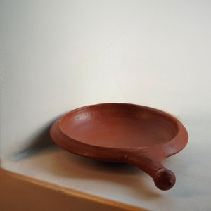 Puede incluir: Un plato de servir de arcilla hecho a mano, de color terracota, con un asa. El plato tiene un cuenco poco profundo y redondo y un asa redondeada con un pequeño pomo en el extremo. El plato está sobre una superficie blanca.