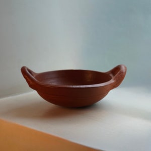 May include: A small, terracotta cooking pot with two handles. The pot is empty and has a reddish-brown color.