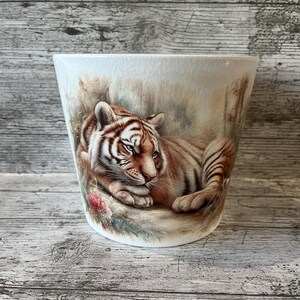 May include: White ceramic plant pot featuring a detailed illustration of a tiger. The tiger is depicted with orange and black stripes, in a relaxed pose. The pot has a flared shape and a painted background.