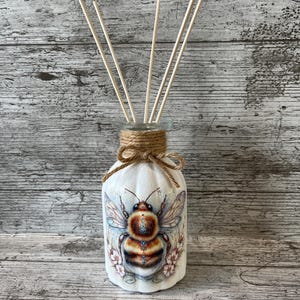 May include: White glass diffuser bottle featuring a detailed bee illustration. The bottle has a jute twine bow around the neck and contains wooden reeds. The bee design incorporates brown, gold, and blue hues, with pink floral accents. A decorative item.