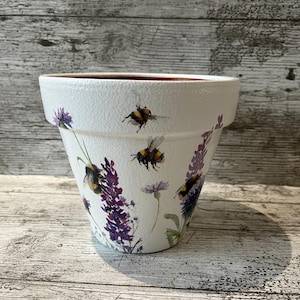 May include: White ceramic plant pot with a textured finish. The pot features illustrations of bumblebees, purple lavender, and wildflowers. The pot has a wide rim and a slightly tapered shape.