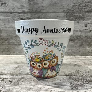 May include: White ceramic flower pot with a painted design of two owls wearing floral hats. The pot has the words "Happy Anniversary" in black script. The owls are surrounded by floral and leaf accents.