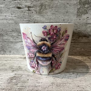 May include: White ceramic plant pot decorated with a detailed illustration of a bumblebee with pink and purple wings, surrounded by pink and purple flowers. The pot has a curved shape and is suitable for indoor or outdoor use.