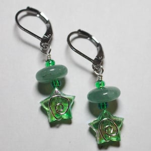 May include: A pair of dangle earrings featuring green star-shaped charms with a silver wire design. Each earring has a green stone and small green beads, suspended from a silver leverback clasp. The earrings have a bright, playful appearance.