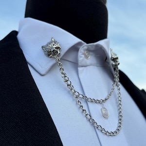 May include: A white dress shirt with a black suit jacket. The shirt has a silver chain collar clip with wolf head accents.