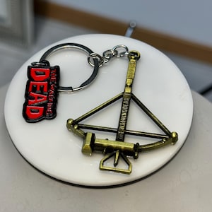 May include: A keychain featuring two charms. One charm is a red and black rectangular tag that reads "DEAD" and "THE WALKING". The other charm is a bronze-colored crossbow with the text "THE WARRIOR".