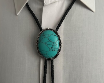 May include: A turquoise bolo tie with a silver-colored oval pendant. The pendant has a textured, marbled appearance and is surrounded by a twisted rope design. The black cord is braided and hangs down over a white collared shirt.
