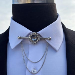 May include: A silver collar pin with a large, faceted, dark gray gemstone in the center. The pin is attached to a double chain with a white button at the end.