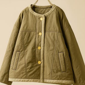 May include: Olive green jacket with a round collar and four gold buttons. The jacket has two front pockets and a light beige trim along the collar and button placket. The jacket is hanging on a beige rope hanger.