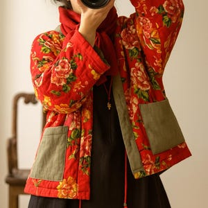 May include: A red floral print jacket with olive green pockets and trim. The jacket features a vibrant pattern of large flowers in shades of yellow, orange, and pink. The jacket is worn over a black dress.