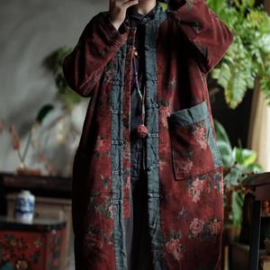 May include: A long, red corduroy coat with floral print accents. The coat features a mandarin collar, a button-up front, and a contrasting teal trim. The coat has two pockets and a decorative tassel. The coat is worn by a person.
