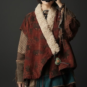 Op de afbeelding: Een persoon draagt een rood jasje met een patroon, een crèmekleurige kraag van imitatiebont en een bijpassende hoed. De jas heeft een bloemenmotief en is gelaagd over een top met patroon. De outfit heeft een bohemien stijl.