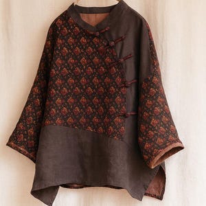 Puede incluir: Una blusa marrón oscuro de manga larga con cuello mandarín y un patrón floral único en la parte superior. La blusa presenta un cierre de botones y un panel marrón liso contrastante en la parte inferior. Las mangas son anchas y la tela parece ser de lino.