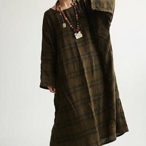 Puede incluir: Vestido largo a cuadros verde oliva con mangas largas y corte holgado. La persona lleva un sombrero beige y un collar de cuentas de madera de varias hebras con colgantes. Una cámara negra se sostiene frente a la cara.