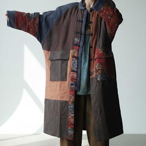 Könnte beinhalten: Ein Patchwork-Mantel in Braun-, Blau- und Rottönen mit floralen Akzenten. Der Mantel hat einen Stehkragen, große Taschen und eine Knopfleiste. Die Ärmel haben ein einzigartiges, weites Design. Der Mantel wird mit einer braunen Hose getragen.