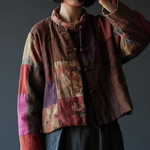 May include: A multi-coloured patchwork jacket in shades of brown, red, and purple. The jacket has a mandarin collar and frog closures. The sleeves are long, and the fabric appears to be a textured cotton or linen blend. The jacket has a vintage aesthetic.