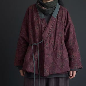 May include: A burgundy floral print jacket with a wrap-style design. The jacket features a gray trim along the collar and sleeves, and a matching tie belt. The jacket is worn over a gray top and striped pants.