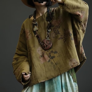 May include: Olive green long-sleeve top with a floral pattern in shades of pink and brown. The top has a loose, flowing fit. A long necklace with a large, round pendant is visible. The bottom of the top is paired with a blue and green skirt.