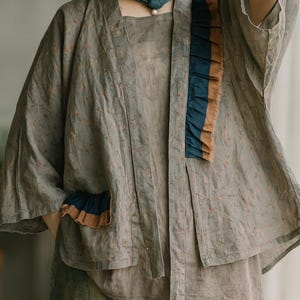 May include: A layered outfit featuring a gray kimono-style jacket with floral patterns, accented with navy and tan ruffles. The outfit includes a matching top and patterned pants. A straw hat and a blue scarf with a pearl pendant complete the look.